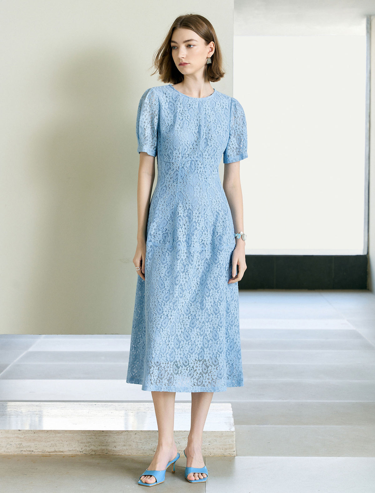 A woman standing in an indoor setting wearing a light blue lace A-line midi dress with short sleeves and knee-length.