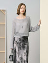 Woman wearing a gray sweater and patterned skirt holding a black handbag indoors.