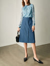 Woman wearing a blue patterned top and blue skirt with black shoes and handbag against a neutral background