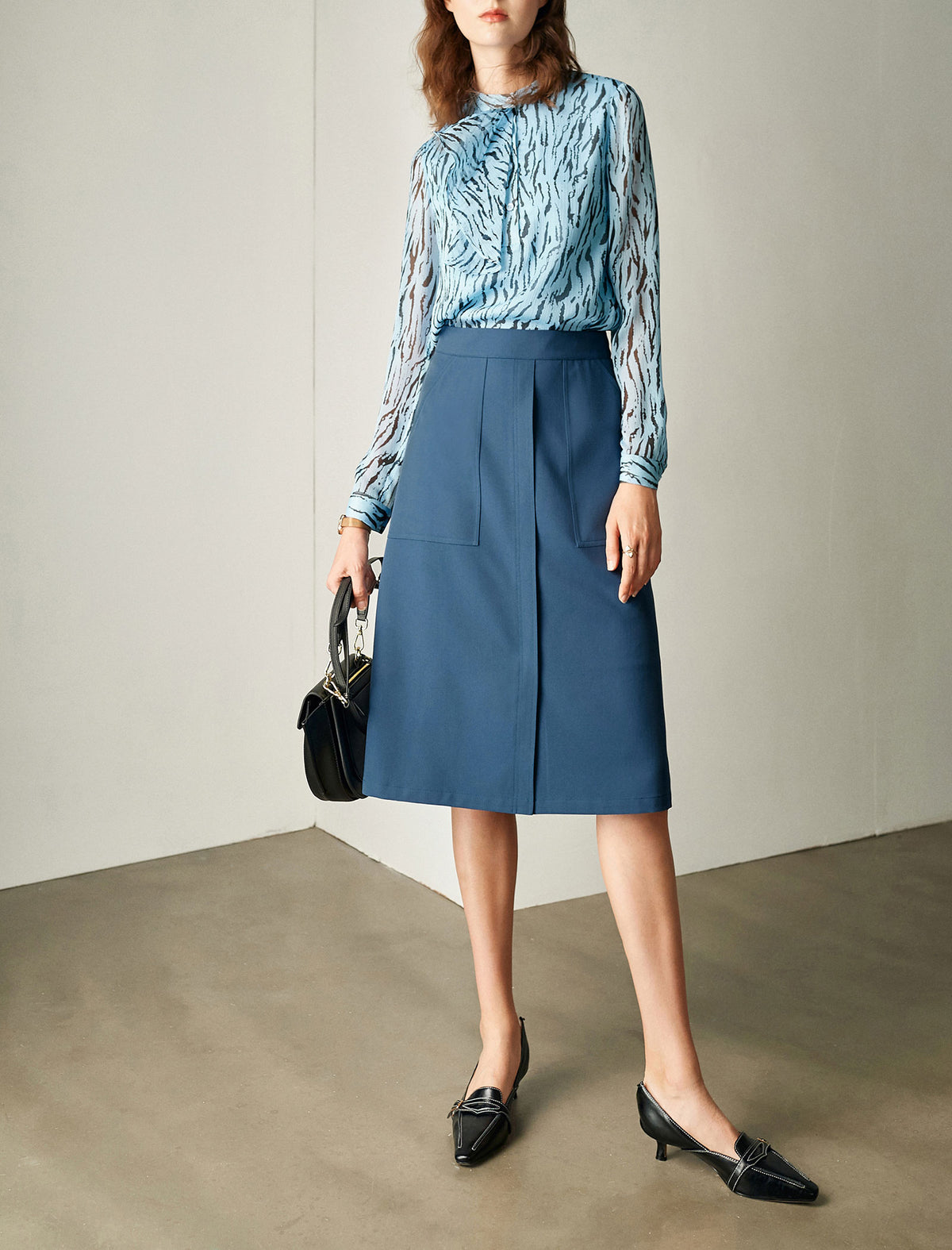 Woman wearing a blue patterned top and blue skirt with black shoes and handbag against a neutral background