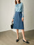 Woman wearing a blue patterned top and blue skirt with black shoes and handbag against a neutral background
