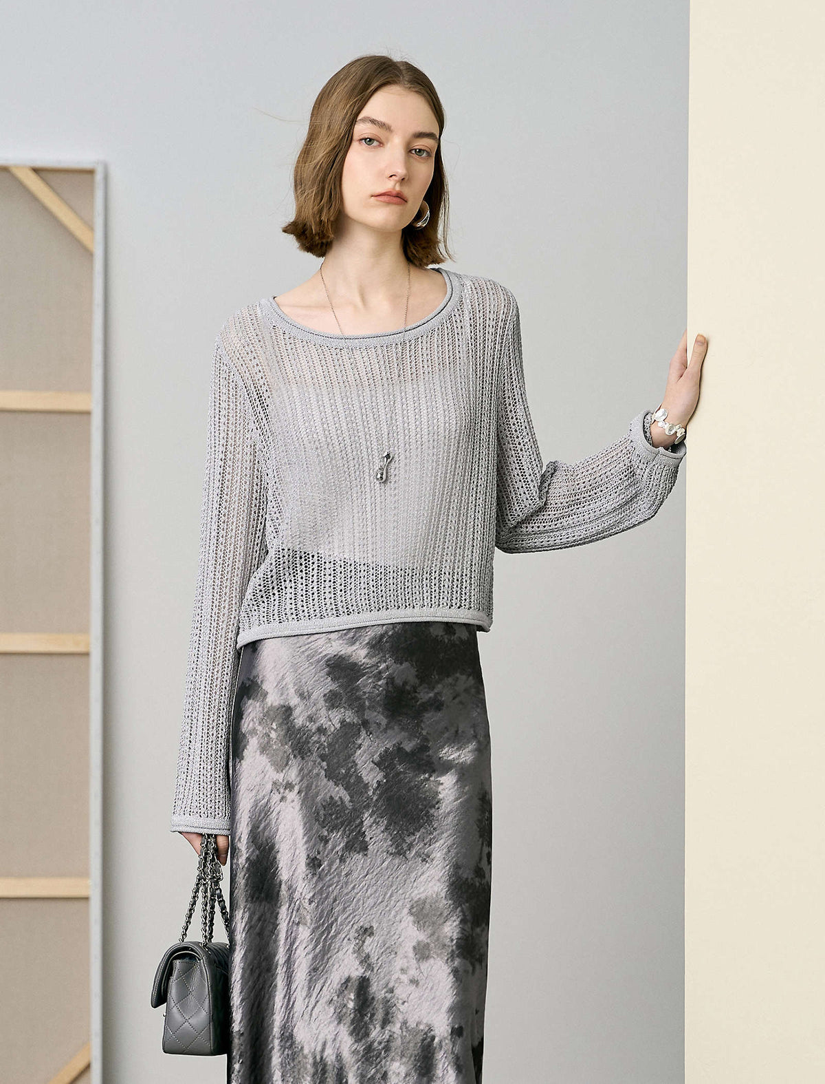 Woman wearing a gray sweater and patterned skirt holding a black handbag indoors.