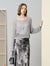 Woman wearing a gray sweater and patterned skirt holding a black handbag indoors.